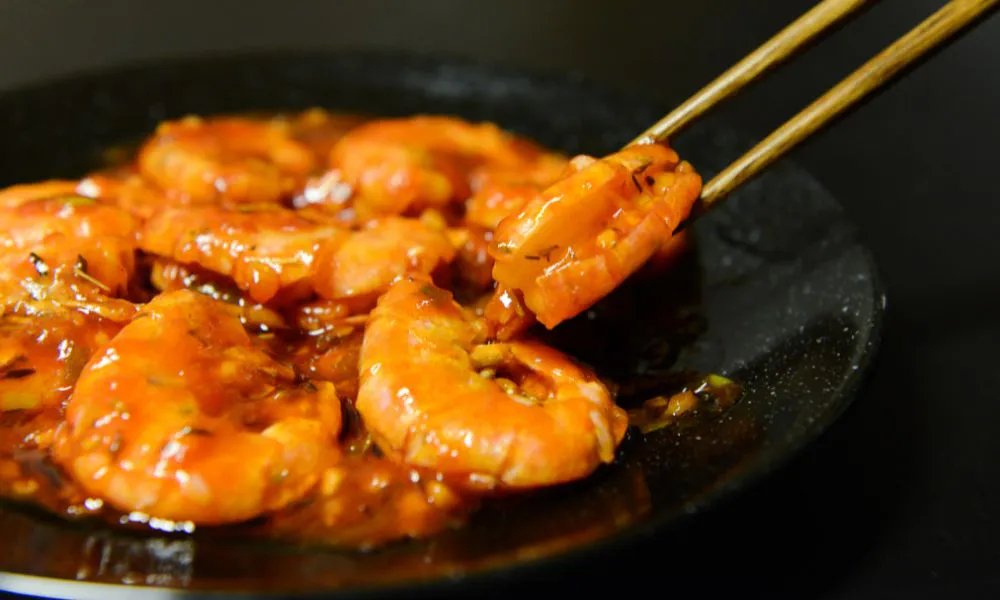 Plump cooked prawns coated in shiny savory red sauce with herbs, served on a dark plate; a single shrimp is picked up by wooden chopsticks for display