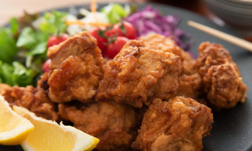 Golden crispy Japanese fried chicken pieces on dark plate, paired with mixed greens, cherry tomatoes, red cabbage and lemon slices for garnish