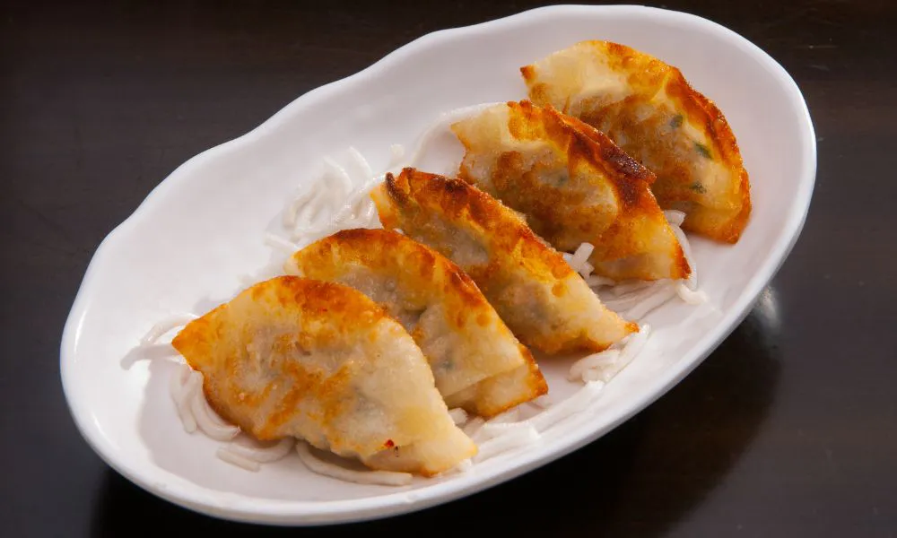 Four potstickers with golden brown crispy crust arranged on a white irregular plate, placed over thin white shredded garnish on dark tabletop