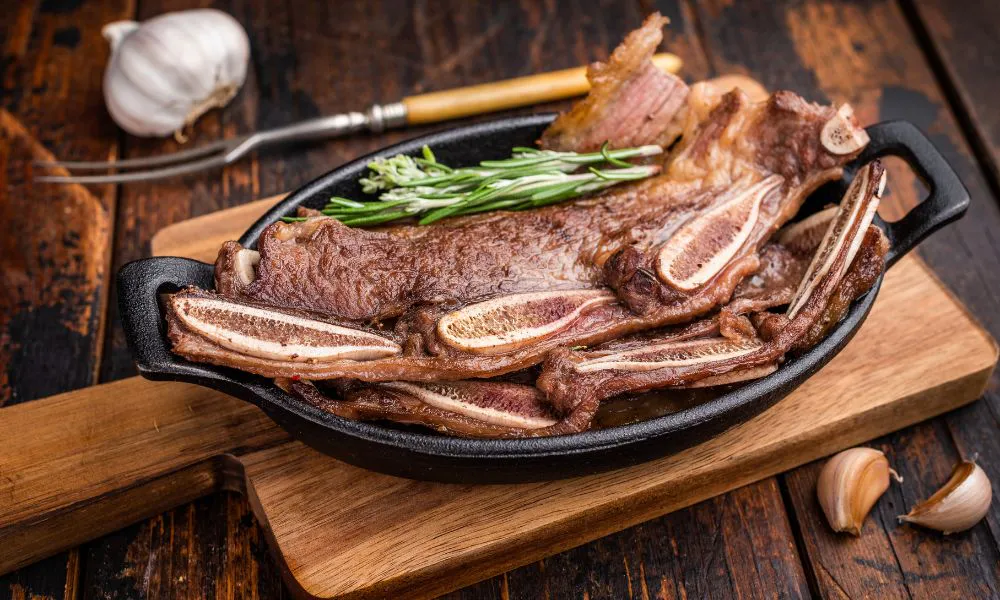 Sliced grilled beef short ribs stacked in a black cast iron skillet, garnished with fresh rosemary, placed on wooden board with garlic cloves nearby