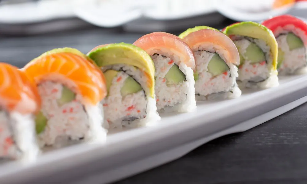 Neatly sliced rainbow sushi rolls lined on white rectangular plate, filled with crab meat and cucumber, topped with colorful raw fish and avocado slices