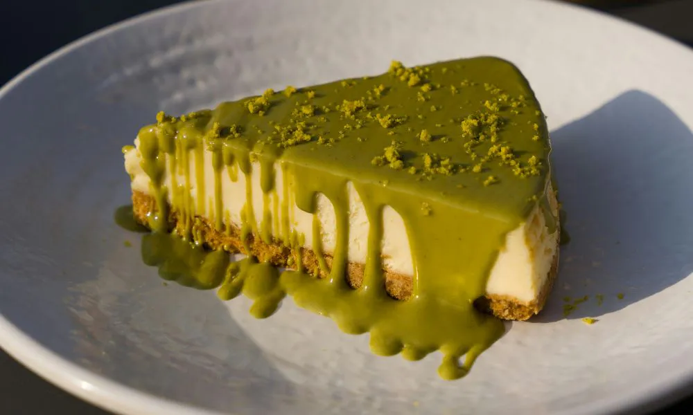 Triangular slice of creamy cheesecake with graham cracker crust, covered in flowing matcha sauce and matcha powder sprinkles on white ceramic plate